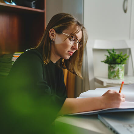 Young woman with glasses writing in a notebook at a desk, with a plant in the background.