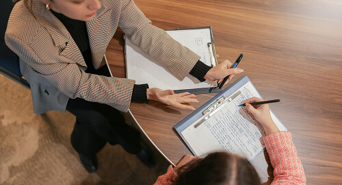 Two people sit at a round table with clipboards and papers. One person gestures while the other writes notes.