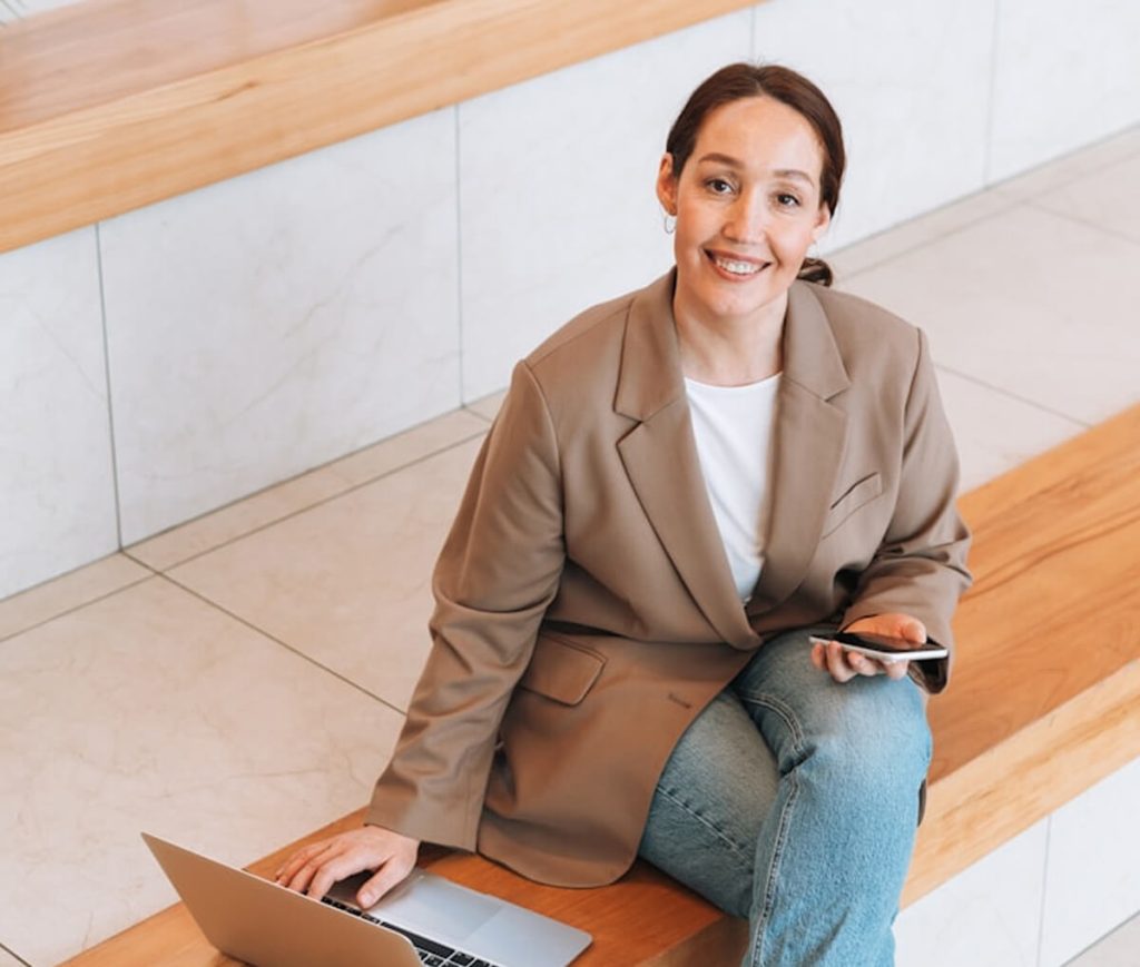 A person in a beige blazer and jeans sits on wooden stairs, holding a smartphone, with a laptop open beside them. They are smiling and looking upward.