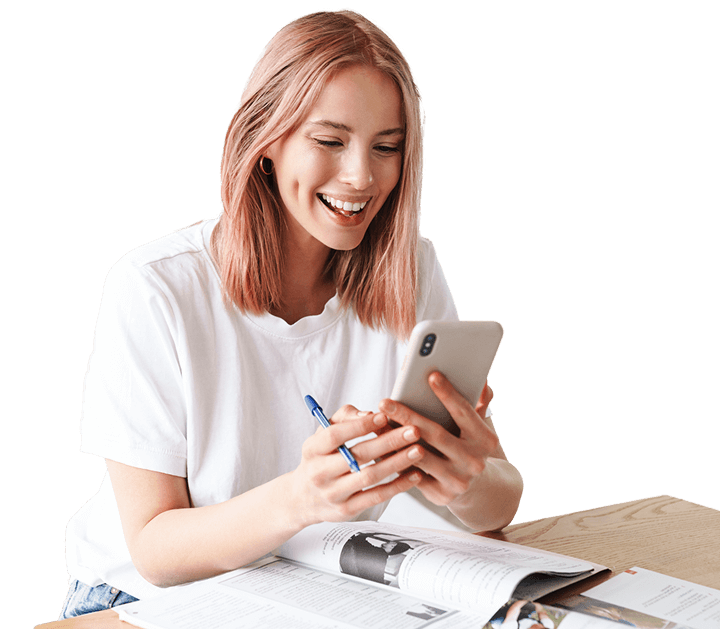 A person with pink hair smiles while looking at a smartphone. They are sitting at a table with an open magazine and holding a pen.