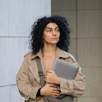 Person with curly hair holding a laptop, wearing a trench coat, standing against a light-colored wall.