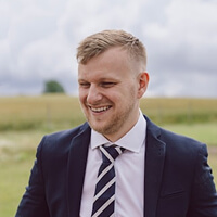 A person in a suit and tie is standing outdoors, smiling, with a blurred grassy field and cloudy sky in the background.