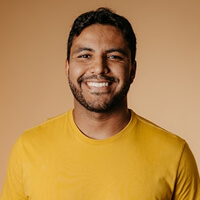 A person smiling, wearing a yellow shirt, stands against a plain beige background.