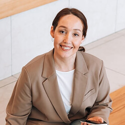 Smiling person with brown hair in a bun, wearing a tan blazer over a white shirt, sits on steps holding a smartphone.